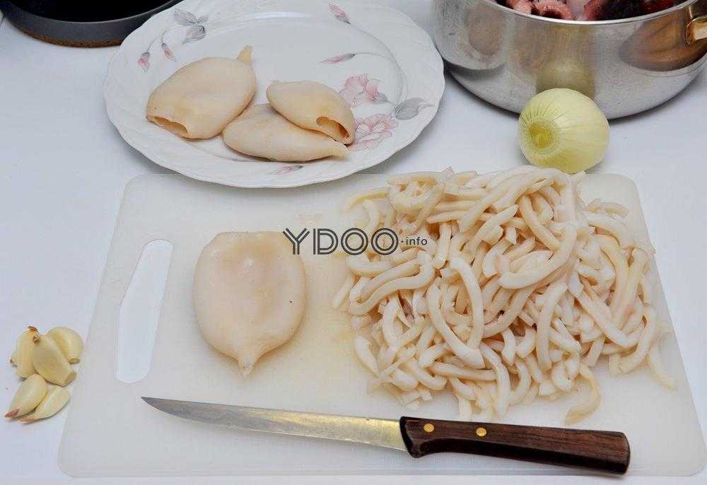 тушки кальмаров а тарелке, нарезанные ножом кальмары на разделочной доске, очищенные зубчики чеснока