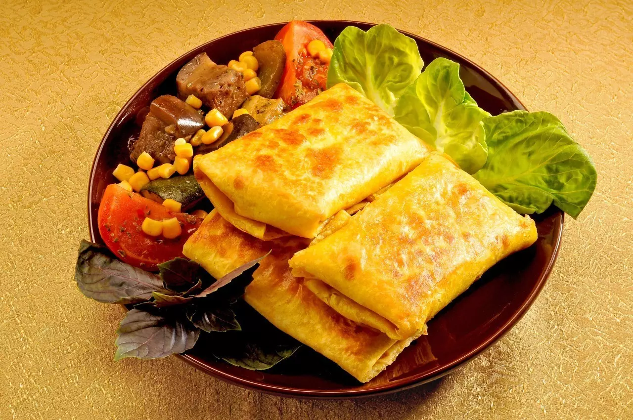 два жареные кусочка чимичанги с листьями салата, кусочками томатов, консервированной кукурузой и листиками базилика на коричневой плоской тарелке на столе