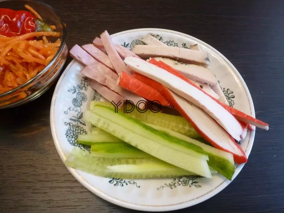нарезанные соломкой свежие огурцы, крабовые палочки и ветчина в белой тарелке на столе, рядом корейская морковь в прозрачной стеклянной миске