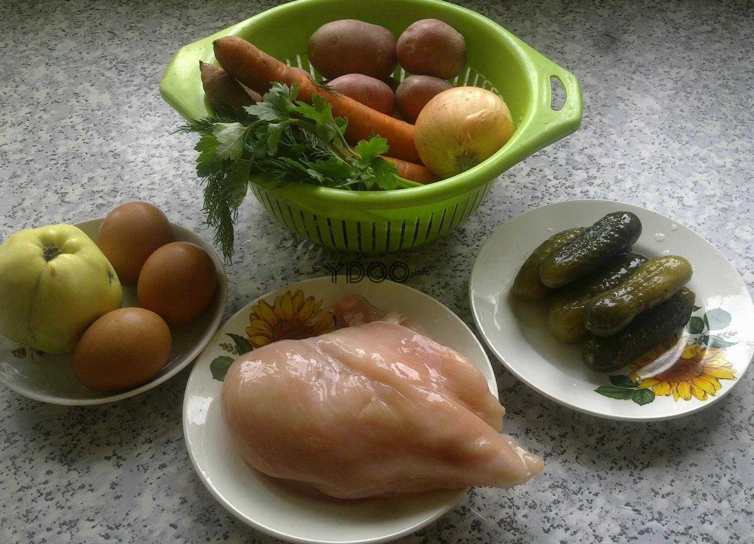 на столе стоит зеленый дуршлаг внутри лежат две морковки, репчатый лук, картофель и зелень, рядом на тарелке лежат соленые огурцы, рядом на тарелке лежит куриная грудка, рядом на блюдце лежит три куриных яйца и яблоко