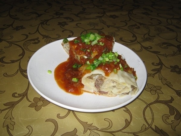 рулет с мясной начинкой внутри, политый томатной подливой и присыпанный рубленой зеленью, на белой тарелке на столе, застеленном скатертью