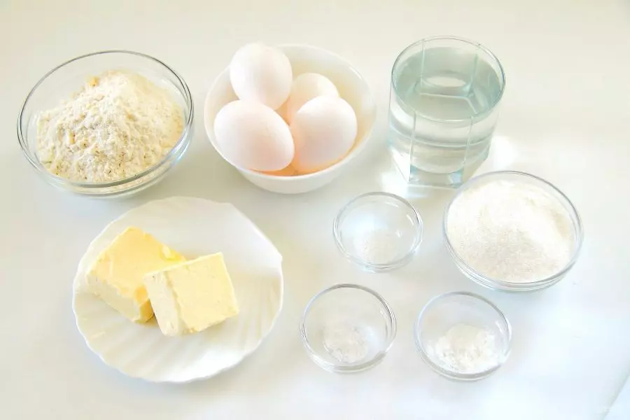 куриные яйца, мука, кусок сливочного масла, соль, сахар и стакан воды на столе
