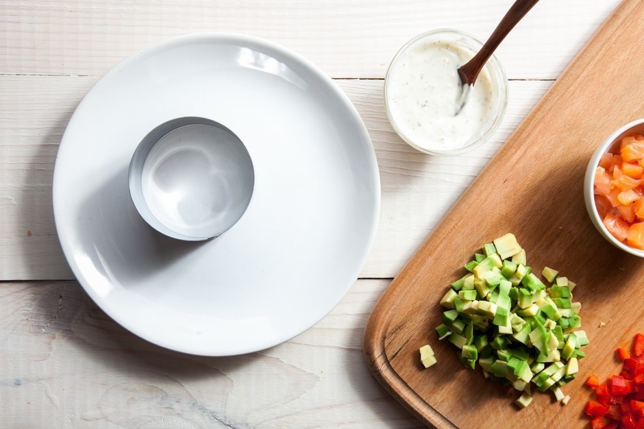 белая плоская тарелка с кулинарным кольцом на столе, рядом нарезанный кубиками авокадо на деревянной разделочной доске и пиала с белым соусом