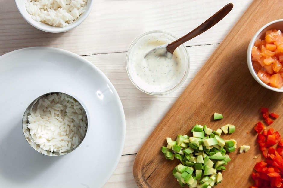 слоеный салат из вареного риса в кулинарном кольце на белой плоской тарелке на столе, рядом пиала с соусом и кубики авокадо на деревянной разделочной доске
