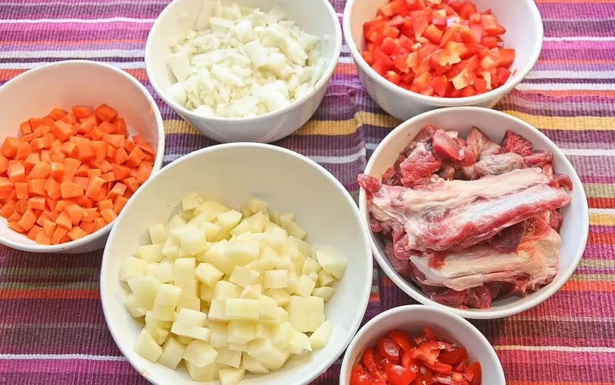 нарезанные кубики картофеля, моркови, репчатого лука, красного сладкого перца, помидоров и мяса в разных белых мисках на столе, покрытом тканью