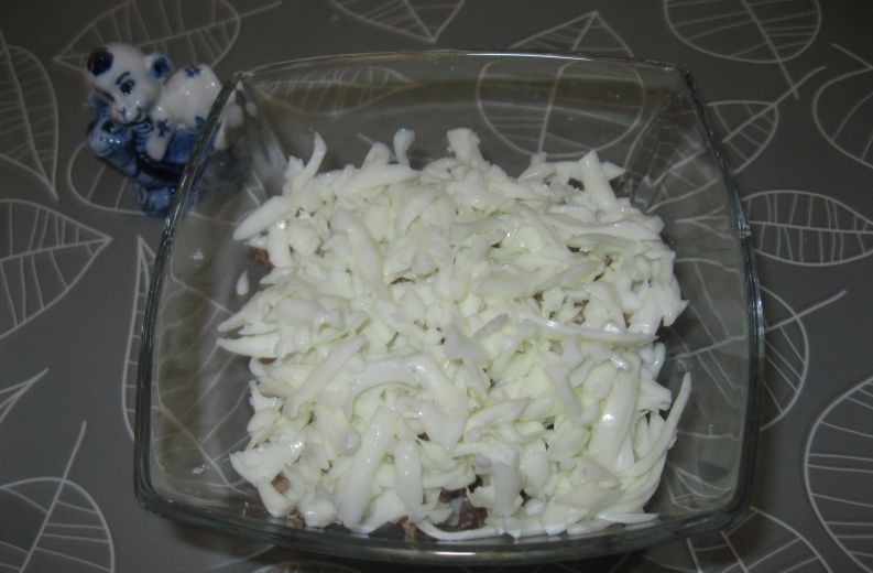 слоеный салат из тертых вареных яичных белков и кусочков консервированного тунца в стеклянной миске на столе