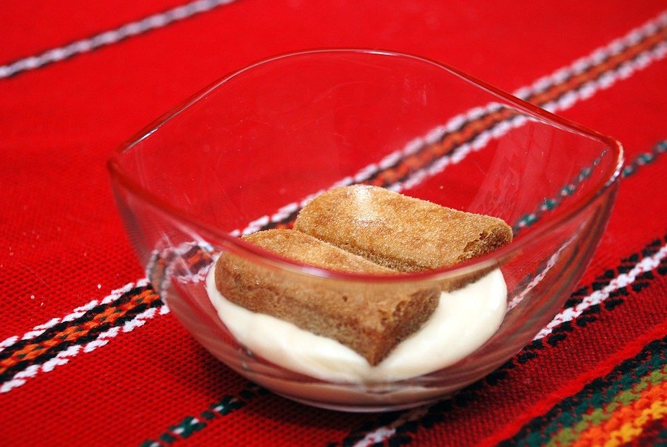 два печенья савоярди, выложенные на сливочный крем, в стеклянной пиале на столе, застеленном скатертью