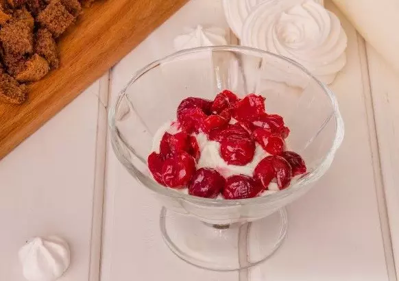 слоеный десерт из творожного крема и вареных ягод вишни в стеклянном стаканчике на столе