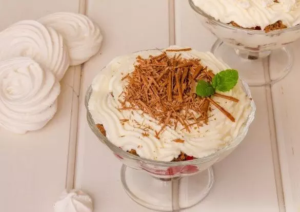 слоеный десерт из творожного крема, присыпанного тертым молочным шоколадом, в стеклянном стаканчике на столе