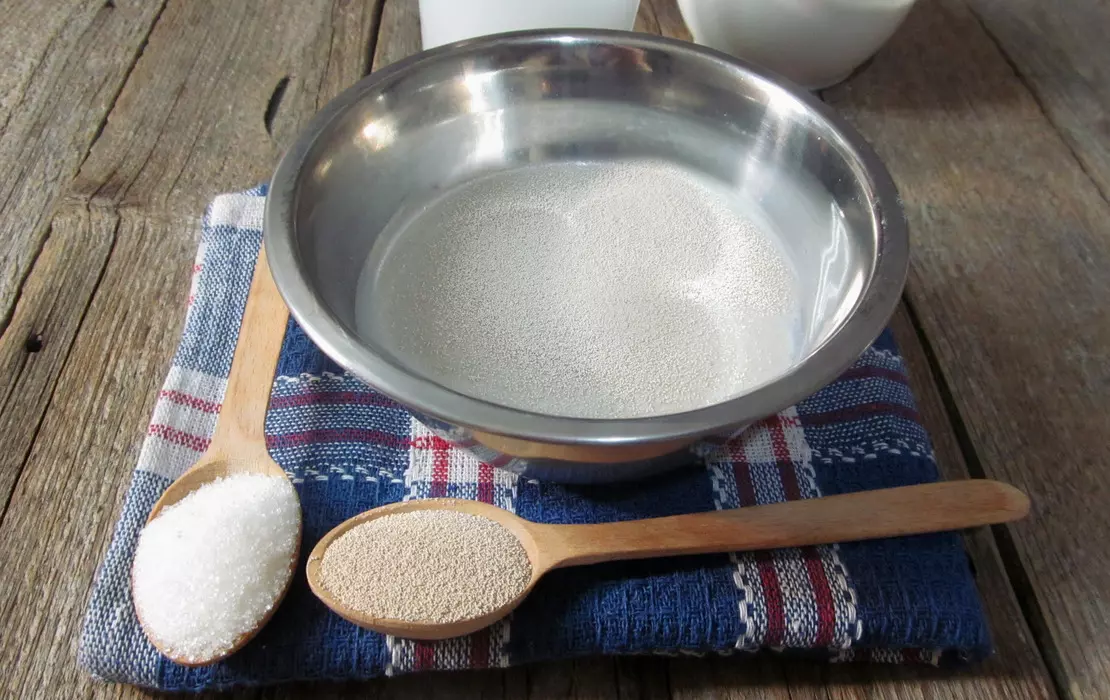 сухие дрожжи и сахар в деревянных ложках, рядом миска с молоком