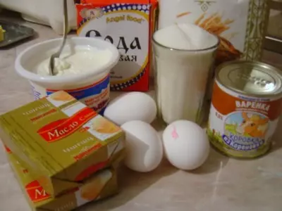 куриные яйца, две пачки сливочного масла, упаковка соды, банка сметаны, стакан сахара, пачка муки и банка вареной сгущенки на столе